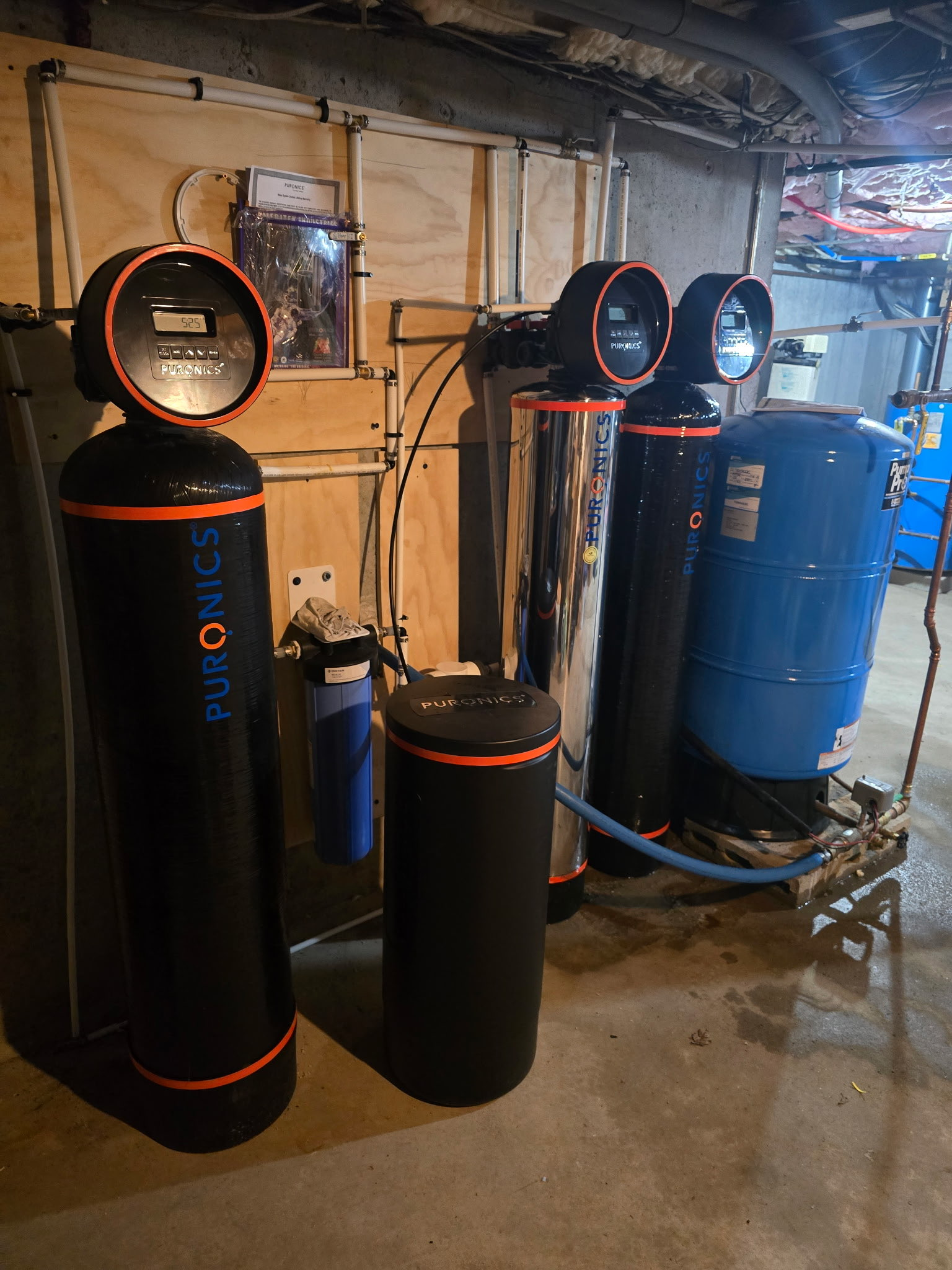Multiple installed Puronics water treatment tanks with orange trim and digital control heads in a residential basement, connected to copper and PVC plumbing.