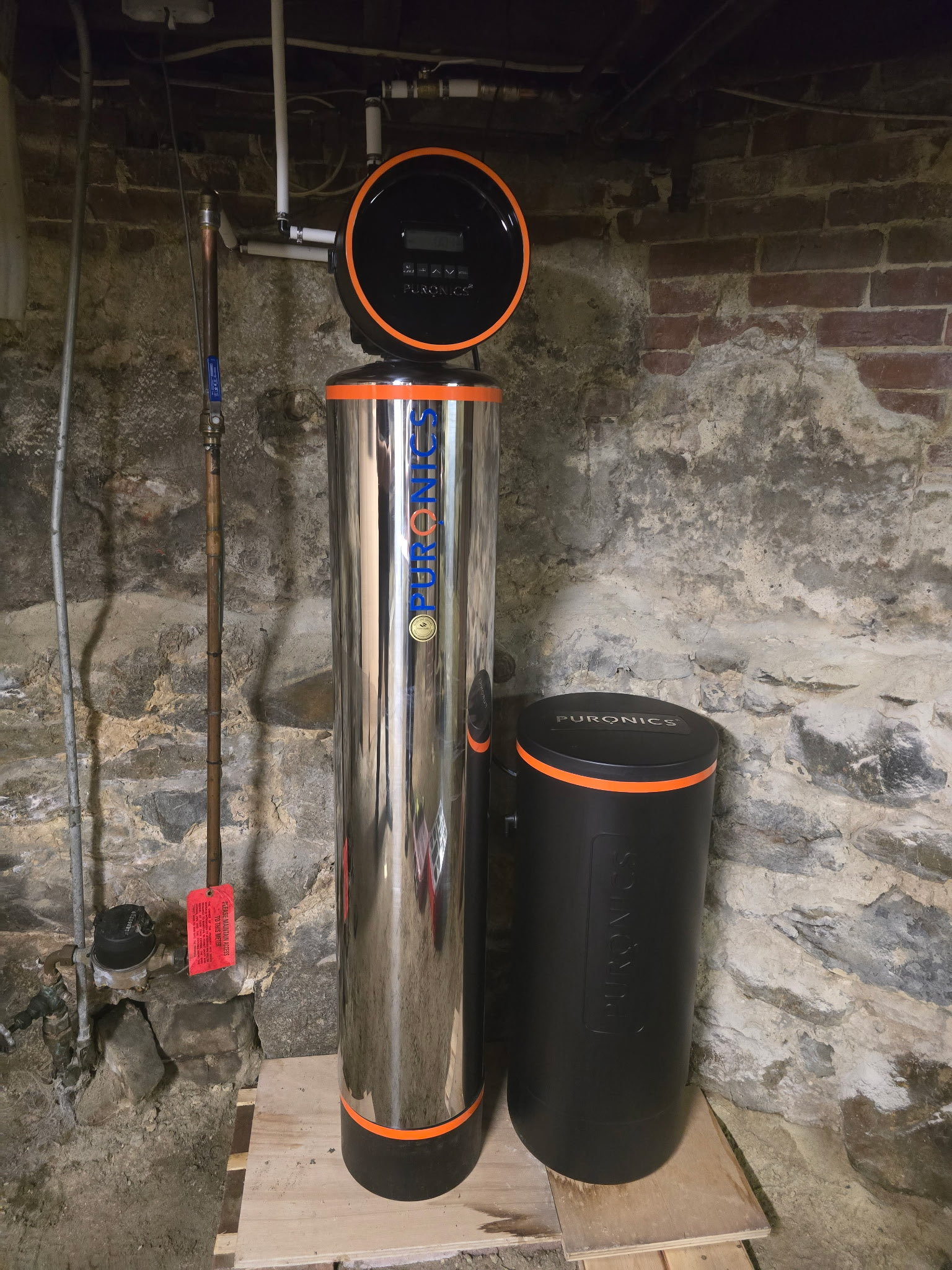 Stainless steel Puronics tank with orange trim and a digital control head installed on a plywood base against a fieldstone basement wall.